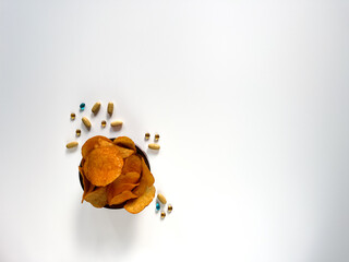 Unhealthy snack food and supplements. A bowl of potato chips next to a scattering of various pills and capsules on a white background. Choosing unhealthy eating over a healthy lifestyle.