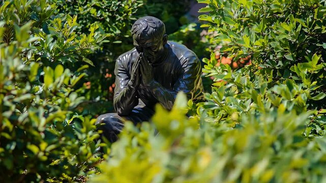 A statue depicts a man in deep thought, surrounded by vibrant greenery in an outdoor setting known for tranquility