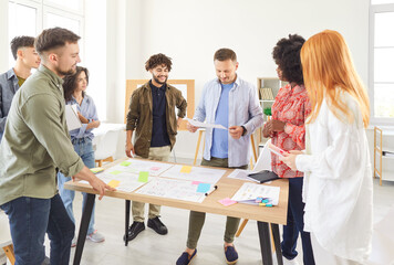 Multiracial group of young people in casual wear working together in collaborative project. Diverse team of students or colleagues standing around desk, brainstorming, discussing ideas, planning work.