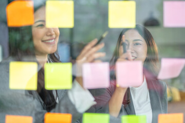 Two Asian businesswomen brainstorming using sticky notes in an office.