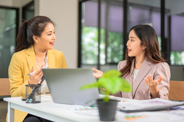  Two professional businesswomen are working on laptop computers sitting at desks in the office. Colleagues are happy and discussing the digital strategy of the Graph