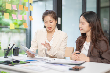  Two professional businesswomen are working on laptop computers sitting at desks in the office. Colleagues are happy and discussing the digital strategy of the Graph