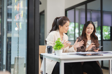 Two professional businesswomen are working on laptop computers sitting at desks in the office. Colleagues are happy and discussing the digital strategy of the Graph