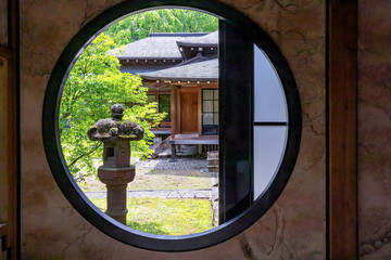 An atmospheric view of a traditional building with a stone lantern and greenery through a round window from Nikko Tammozawa Imperial Villa, Japan