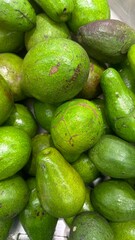 The image shows a large pile of fresh, green avocados, some with darker blemishes, indicating a natural appearance and varying stages of ripeness