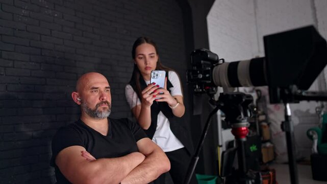 Bearded man sits with hands crossed on his chest behind the camera. Lady standing beside taking video on smartphone.