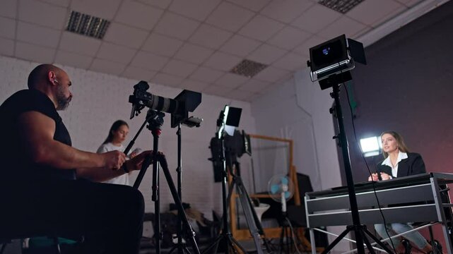 Bearded cameraman sits behind the camera recording a woman at desk. Low angle view. Girl with phone in hands stands at backdrop.