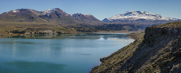 Turquoise waters of Lake Caviahue under rocky landscape and snowcapped Copahue volcano. Caviahue, Neuquen, Argentina