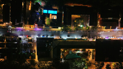 Aerial view captures the vibrant energy of a city street at night, with buildings, pools, and parked cars bathed in the warm glow of artificial lights, creating a captivating urban nightscape