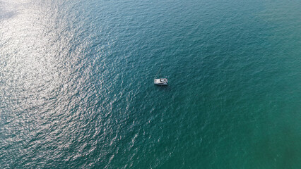 Aerial view of a luxurious catamaran sailing on calm turquoise water with sunlight reflecting on the surface, creating a mesmerizing visual contrast between the white vessel and the deep blue sea