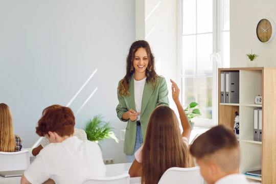 School happy teacher and students learning in classroom, asking pupils at desk. Female tutor, schoolchildren group during lesson at school, student girl raises hand, ready to answer questions in class