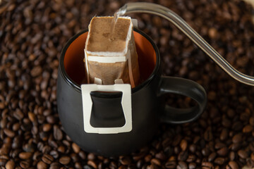Close-up view of hot water pouring from metal kettle into white paper drip coffee bag with ground arabica hanging on black and orange mug standing on roasted coffee beans.