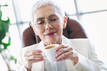 Mature businesswoman enjoying coffee in office chair with closed eyes showing elegant style and...