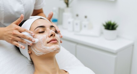 Professional beautician applying white facial cream to woman with closed eyes in modern spa salon. Specialist performing relaxing skincare treatment with moisturizing mask. Beauty therapy