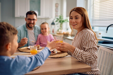 Happy mother giving her son glass of orange juice at dining table.