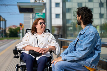 Naklejka premium Disabled woman in wheelchair smiling and talking with man at tram station - wheelchair and disability concept