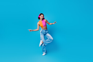 Obraz premium Stylish young woman in colorful summer outfit dancing and enjoying music with headphones on vibrant blue background