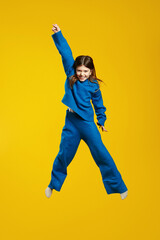 Vertical photo of energetic kid in a blue outfit joyfully leaps with a big smile and raised hand on...
