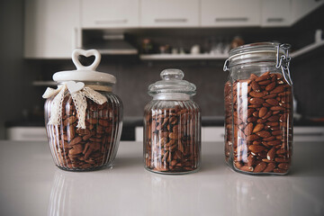 Three glass jars filled with almonds sit neatly on a kitchen counter. The jars have decorative lids, adding a touch of elegance to the home kitchen setting. Healthy food and lifestyle concept