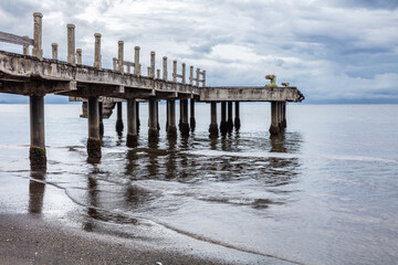 Pier in Lovina Beach, Northern Bali, Indonesia. Calm water; beach in foreground. Cloudy sky overhead. 
