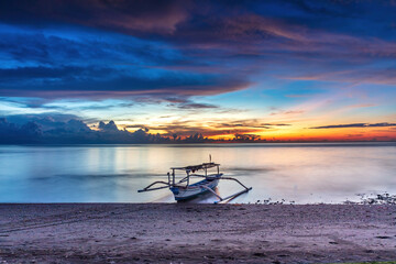 Ocean sunset at Lovina Beach, northern Bali, Indonesia. Beach with boat in theforeground; setting sun surrounded by colorful clouds. Sun's reflection on the water. 
