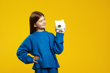 Cheerful kid in a cozy blue sweater admiring her piggy bank, exuding a sense of savings and financial awareness, isolated against yellow background with free empty space.