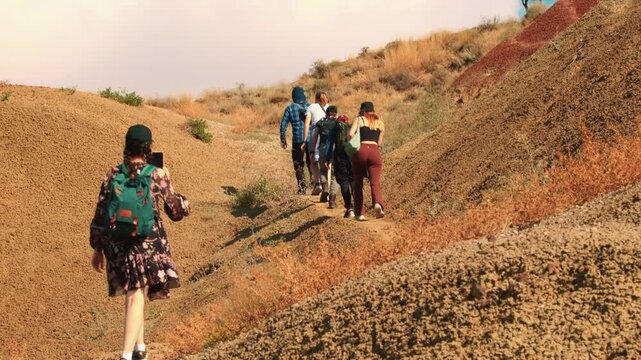 Multicultural young hikers climbing sandy slopes during guided tour in desert highlands, exploring extreme terrain and bonding through outdoor travel experience. Back view.