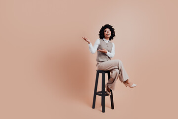Confident businesswoman seated on a stool gesturing with elegance against a beige background,...