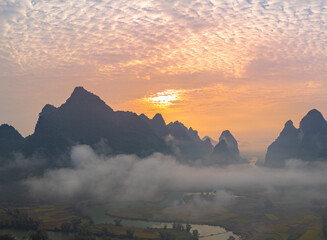 A breathtaking aerial view of a dramatic sunrise illuminating the sky with vibrant orange and red hues over mist-covered karst mountains. The serene landscape, dotted with valleys and rivers,