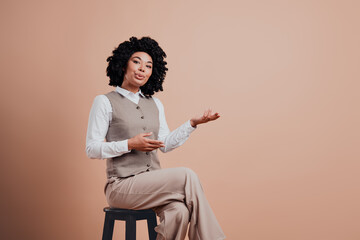 Confident Businesswoman Gesturing While Sitting on Stool in Stylish Formalwear Against Beige...