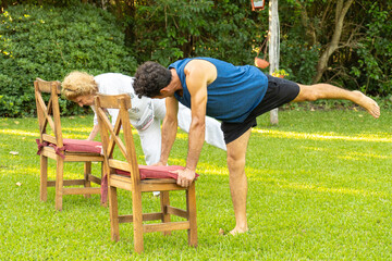 Senior couple doing balancing yoga pose in garden using chairs