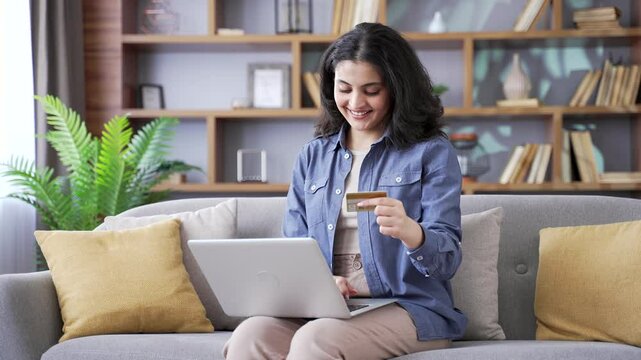 Happy young woman doing online shopping entering credit card data on a laptop computer sitting on sofa in the living room at home. Smiling female happily makes a purchase, enjoy e-commerce e-shopping