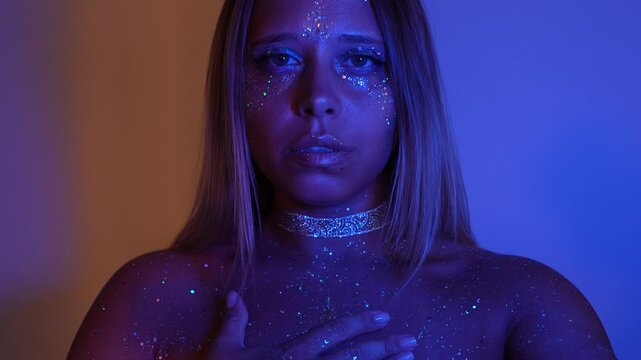 A young mesmerising cosmic woman opens her eyes and smiles gently, hand resting on her heart. A deep breath awakens the memory of her divine essence, glowing in blue and sunset light. The alien soul