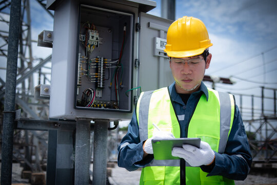 Power station engineer working outdoors with safety helmet and vest checking electrical control panel and taking notes on digital tablet in industrial environment