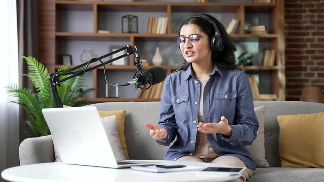 Confident young woman recording podcast using laptop computer and microphone in home office. Female vlogger influencer conducts online broadcast, interview or webinar, speaks remotely via video call