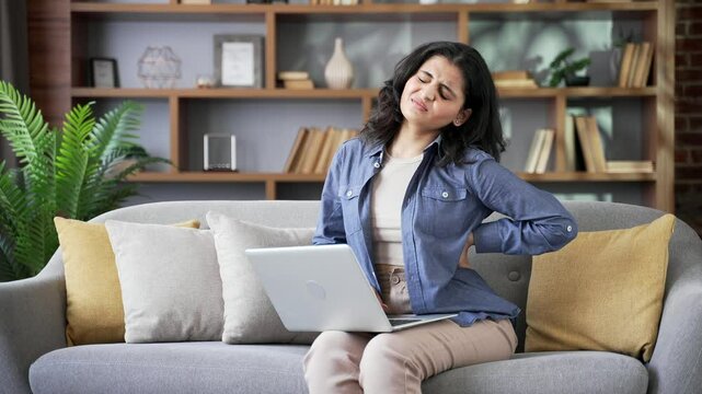 Tired young woman suffering from back pain while working on a laptop sitting on sofa in living room at home. Upset exhausted female massages painful lower back muscles. Discomfort due to poor posture
