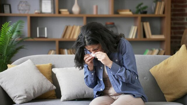 Allergic young woman sneezes and wipes her nose with handkerchief. Unhealthy adult female suffering from seasonal allergies sitting on sofa in living room at home. Girl is sick with cold, virus or flu