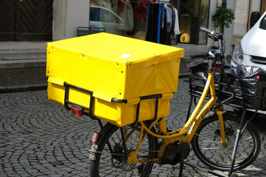 Electric cargo bike with a bright yellow delivery box, used for urban postal services, seen on a historic European cobblestone street.