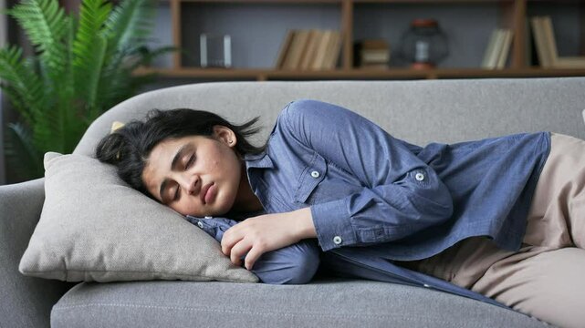 Exhausted young woman tired after hard working day lies down on sofa in living room at home. Overworked female brunette wants to sleep, puts her head on the pillow and closes her eyes, falls asleep