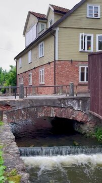 Small waterfall flows beneath old red brick bridge and traditional wooden house on a cloudy day
