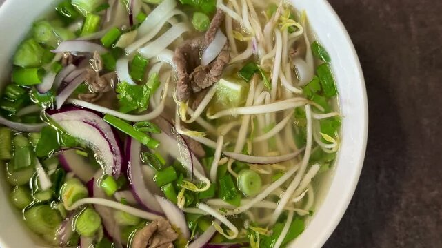 A plate of Vietnamese beef noodle soup "Pho Bo", consisting of broth, rice noodles, herbs and thin slices of meat, on the table in the restaurant. This makes the meat tender and juicy. Close-up
