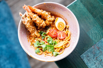 A bowl of ramen with tempura shrimp, half a boiled egg, cherry tomatoes, cilantro, and noodles in a rich broth, served on a green table. Vibrant colors, fresh ingredients, and appetizing presentation