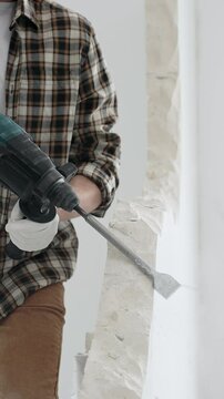 Unrecognizable male construction worker wearing beige checkered shirt and protective gloves, is demolishing white wall with rotary hammer drill, generating dust, close up view. Renovation concept