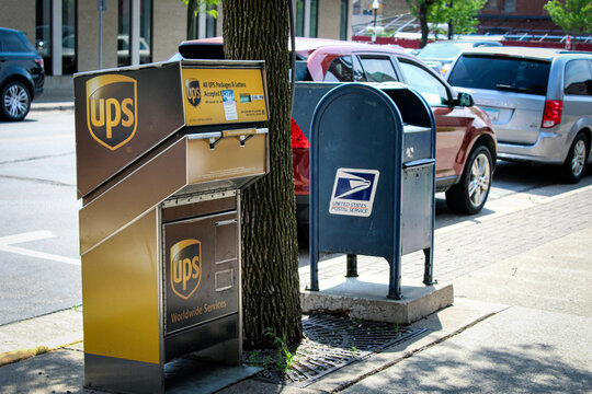 A UPS and USPS drop-off box next to each other on the sidewalk with cars on the street in the background. (Anoka, Minnesota - June 2, 2025)