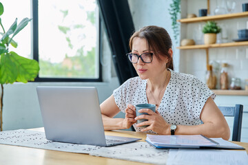 Middle aged serious woman working with laptop sitting at table at home