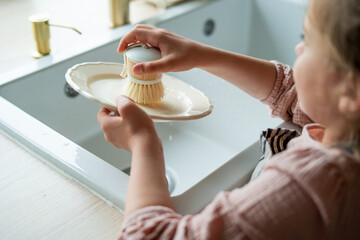 Closeup of children's hands washing a plate near the sink in the kitchen. Eco dishwashing brush.