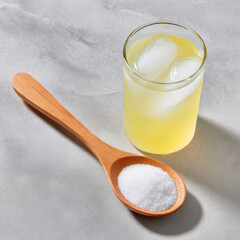 Wooden spoon filled with white powder is placed next to refreshing glass of iced lemon drink on light gray surface