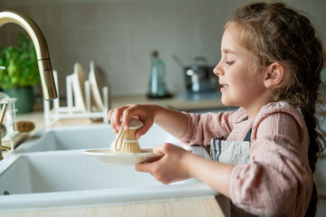 little girl of 7 years old washes a plate in the kitchen near the sink. Child cleans the dishes....