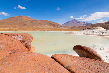 Striking red rocks frame a serene turquoise lake in Chile's Atacama, with majestic volcanoes under...