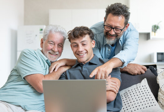 Happy multigenerational family sitting on sofa using laptop together - grandfather with mature son and young grandson enjoying carefree moments together learning use of computer